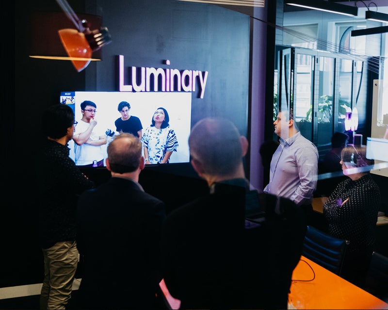 People in a meeting room standing round a TV with other people in another meeting room on it