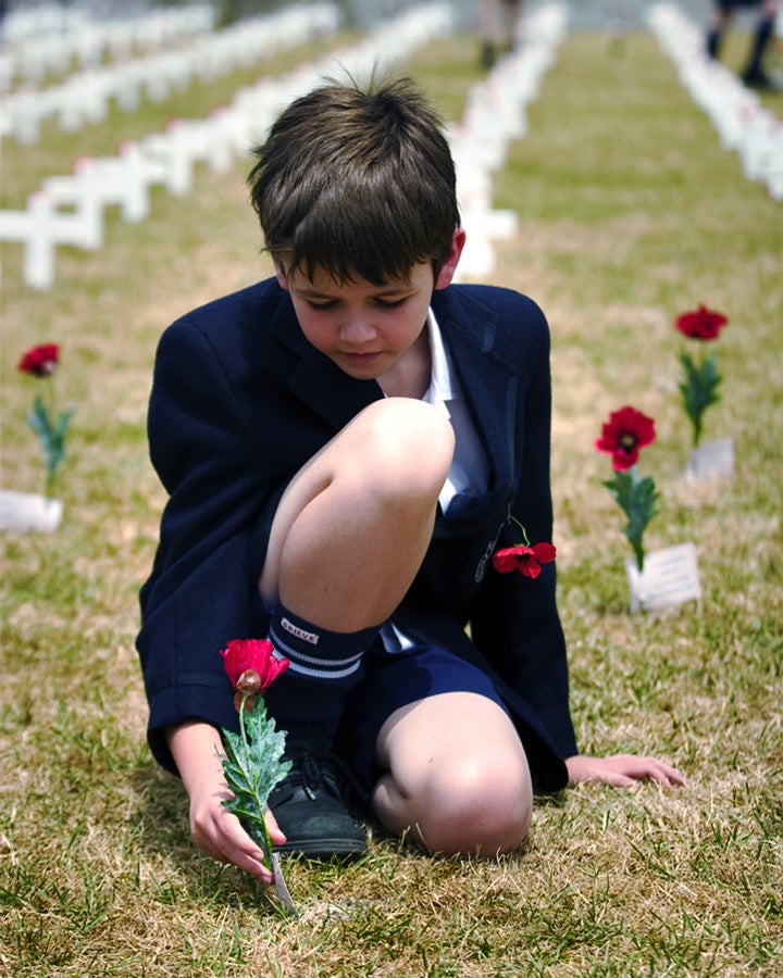 Boy in school uniform crouched on lawn sticking a poppy in the ground