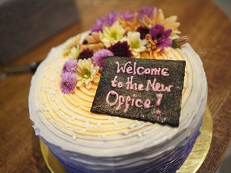 Round white cake with purple and yellow flowers and a plaque that says "Welcome to the new office!"